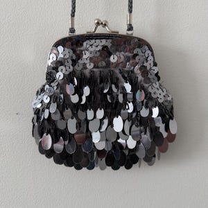 Peut inclure: Un petit sac à main argenté à paillettes avec une fermeture à fermoir et une bandoulière grise tressée. Le sac est recouvert de paillettes ovales qui se chevauchent et pendent du bord inférieur. Le fermoir est argenté et la bandoulière est grise.