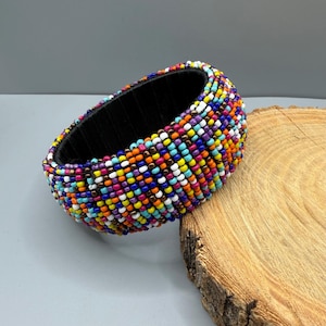 May include: A wide cuff bracelet featuring a dense, multi-coloured pattern of small glass beads in shades of blue, orange, yellow, white, purple, and pink. The interior of the bracelet is lined with a smooth, solid black material.