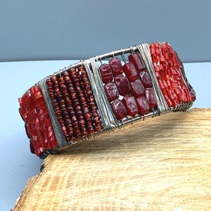 May include: A handmade bracelet featuring a silver wire frame with sections of red beads. The bracelet has a rectangular shape with alternating sections of small, round beads and larger, rectangular beads. The beads are a deep, rich red color.
