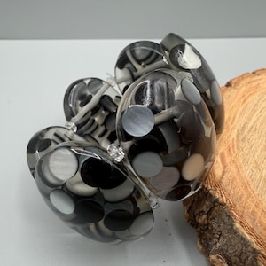 Puede incluir: Una pulsera elástica gruesa compuesta por múltiples eslabones de resina redondeados y translúcidos. Cada eslabón presenta un diseño en capas con discos circulares incrustados en varios tonos de negro, blanco y gris, creando un efecto monocromático.