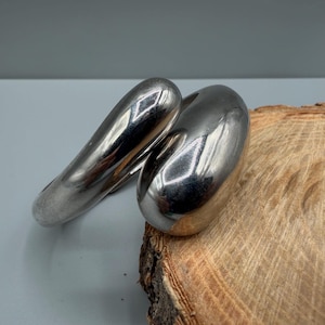 May include: A polished silver-tone metal cuff bracelet with a smooth, rounded, and organic shape. The bracelet features a continuous, thick band that curves into a sculptural form, reflecting light across its highly reflective, mirror-like surface.