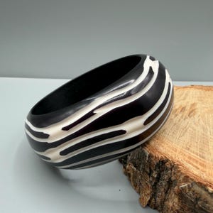 May include: A thick, circular bangle bracelet featuring a high-gloss black finish with irregular, flowing white stripes that wrap around the exterior surface. The bracelet has a smooth, polished texture and a wide, substantial profile.