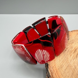 May include: A stretch bracelet composed of multiple rectangular segments in vibrant red and deep black. The segments feature a glossy finish with a white floral pattern on the front piece, connected by elastic cord to form a flexible circular band.