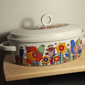 May include: White oval casserole dish with lid. The dish features a colourful floral and bird design in red, orange, green, and blue. It has two handles and a loop handle on the lid. The dish is sitting on a wooden surface.