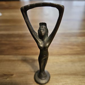 May include: A bronze-colored figurine of a woman with arms raised, holding a curved frame above her head. The figure has a detailed face and is standing on a circular base. The statue is on a wooden surface.