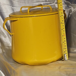 Puede incluir: Una olla de esmalte amarillo brillante con tapa y dos asas laterales, más un asa en la tapa. La olla mide aproximadamente 20 cm de alto y 20 cm de diámetro. Una cinta métrica está al lado de la olla.