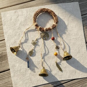 May include: A circular woven twig wreath adorned with hanging jute strings. The strings feature three brass bells, a metal tree of life charm, a star charm, a leaf charm, a small blue crystal vial, a red bead, a yellow bead, and a feather charm.