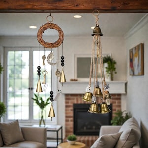 May include: Two hanging wind chimes. One features a woven wreath with hanging keys, crescent moon, and brass bells. The other has multiple brass bells suspended from a rope. The background shows a living room with a fireplace and windows.