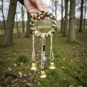May include: A circular wreath made from woven twigs, green leaves, and twine. It features three gold-coloured bells, a pentagram charm, black beads, and a purple crystal. The wreath is held up against a blurred outdoor background of trees and grass.