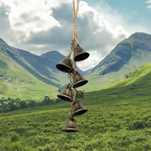 Puede incluir: Un carillón de viento decorativo con seis campanas de color bronce suspendidas de una cuerda natural. Las campanas están dispuestas en cascada, con detalles decorativos de metal. El fondo muestra una vista panorámica de colinas verdes y un cielo nublado.