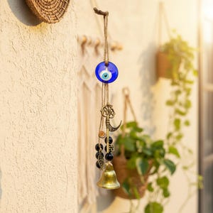 May include: A wall hanging ornament with a blue glass evil eye, a decorative key, a crescent moon, black beads, and a small brass bell. The charm is suspended from a natural twine cord.