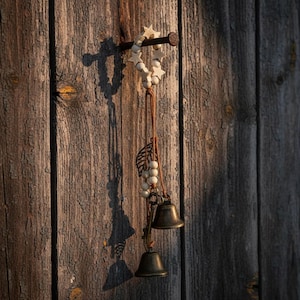 Puede incluir: Un carillón rústico con una guirnalda de estrellas y cuentas de madera, una hoja de metal y dos campanas de bronce. El carillón cuelga de un clavo en una pared de madera desgastada, proyectando una larga sombra a la luz del sol.