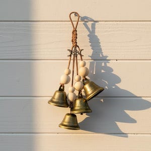 May include: A decorative wind chime with five antique brass bells and wooden beads. The chime is suspended from a braided brown cord with a metal charm. The bells are different sizes and hang at varying lengths.