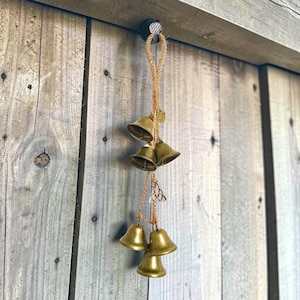 Op de afbeelding: Een hangende windgong met zes messing klokken van verschillende maten. De klokken zijn opgehangen aan een natuurlijk touw en versierd met een kleine bedel en een Keltische knoop. De windgong hangt tegen een verweerde houten achtergrond.