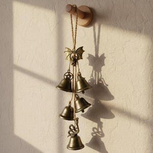 May include: A bronze-coloured wind chime with five bells hanging from a wooden mount. The chime features a bat-shaped top, a star pendant, and a snake-shaped bell at the bottom. The bells are suspended by a natural rope, creating a decorative piece.