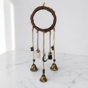 Op de afbeelding: Een handgemaakte dromenvanger met een geweven takkenring. Het heeft hangende jute koorden, kleine glazen flesjes, messing bellen en een maansikkelsieraad. Ontworpen voor woondecoratie.