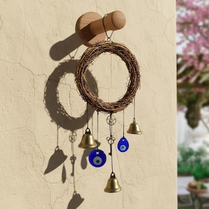Puede incluir: Un carillón decorativo con una corona tejida, campanas de latón, amuletos de ojo turco azules y un colgante de llave. Cuelga de un gancho de madera en una pared texturizada. Diseño natural y metálico.