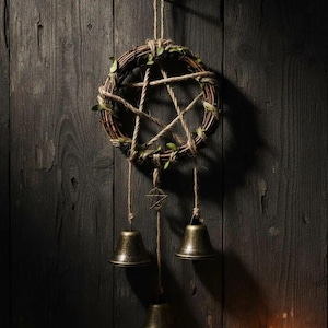 May include: A rustic wall hanging featuring a woven wreath with a pentagram design made of twine. The wreath is adorned with small green leaves and hangs from a jute rope. Three bronze bells dangle below, adding a touch of charm.