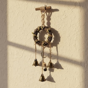 May include: A rustic wall hanging featuring a woven wreath with small green leaves and twine accents. Below the wreath hang three small brass bells and a crescent moon charm. The piece is suspended from a wooden dowel with a knotted rope.