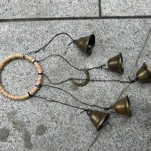 Puede incluir: Un carillón decorativo con un anillo de yute tejido y cuatro campanas de color bronce suspendidas por cuerdas negras. Un pequeño dije en forma de media luna cuelga del anillo. Las campanas están dispuestas a diferentes longitudes.