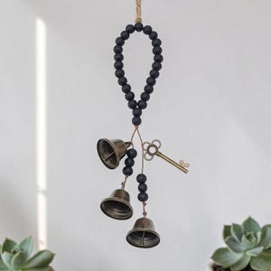May include: A decorative hanging ornament featuring three bronze-colored bells, black wooden beads, and a vintage-style key. The ornament is suspended from a natural rope, creating a rustic and charming aesthetic.