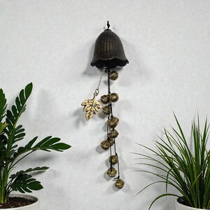 May include: A bronze-colored wind chime with a bell-shaped top and a series of small, round bells hanging below. A decorative leaf is attached to the chime with a chain. The wind chime is hanging on a white wall.