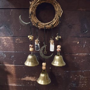 May include: A rustic wall hanging featuring a woven wreath with hanging charms. The charms include small bells, glass vials, wooden beads, and a crescent moon pendant. The color palette is warm, with bronze, brown, and natural tones.