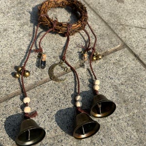 Peut inclure: Un carillon rustique avec une couronne tressée, des cloches en bronze suspendues, de petits flacons en verre, des perles en bois et un charme de croissant de lune. Le carillon est suspendu par des cordons bruns, créant une esthétique naturelle et bohème.