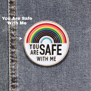 May include: A round enamel pin with a rainbow design and the words "YOU ARE SAFE WITH ME" in black lettering. The pin has a white border and is set against a denim background.