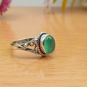 May include: A silver ring with an oval, light green gemstone. The ring has a decorative design with a twisted rope border around the stone and ornate swirls on the band. The ring is set on a light brown surface.
