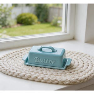 Könnte beinhalten: Hellblaue Keramik-Butterdose mit Deckel und Griff, auf einer cremefarbenen, ovalen, gewebten Matte. Das Wort "Butter" ist auf der Vorderseite eingeprägt. Im Hintergrund ist ein Fenster und ein Garten zu sehen.