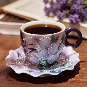 Peut inclure: Une tasse et une soucoupe à fleurs remplies de liquide foncé. La tasse et la soucoupe présentent un motif de tulipes dans des tons violets et lavande. La tasse a une anse violette et est posée sur une surface en bois.