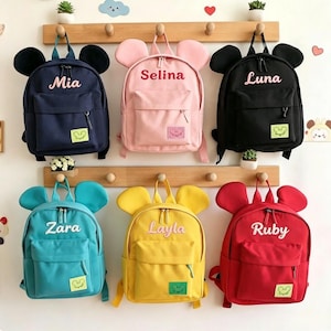 Peut inclure: Six sacs à dos avec des oreilles de souris, chacun doté d'une poche avant, de fermetures éclair et d'un prénom brodé en blanc ou en rouge. Les sacs sont bleu marine, rose, noir, bleu sarcelle, jaune et rouge, avec un petit écusson souris.