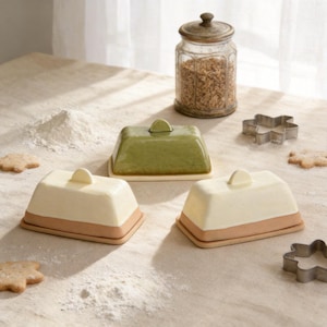 Könnte beinhalten: Drei Keramik-Butterdosen, zwei in Creme und Braun, eine in Grün, stehen auf einer leicht bemehlten Oberfläche. Ein Glas mit Deckel und ein paar Ausstecher sind ebenfalls vorhanden. Die Butterdosen haben eine rechteckige Form mit Deckel und kleinem Griff.