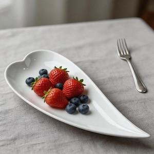May include: A white, heart-shaped serving dish filled with fresh strawberries and blueberries. A silver fork rests on the gray tablecloth next to the dish. The bright red strawberries contrast with the dark blue blueberries.