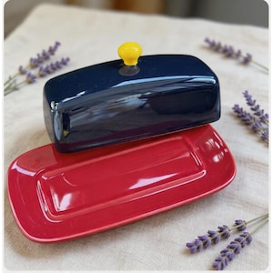 May include: A rectangular red ceramic butter dish with a navy blue lid. The lid has a small yellow knob. The dish is sitting on a neutral-colored surface, with sprigs of lavender in the background.
