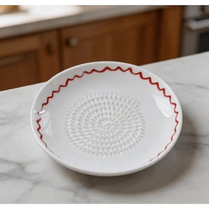 May include: White ceramic grater plate with a red wavy rim. The center of the plate features a textured surface for grating. This kitchenware item is designed for food preparation.
