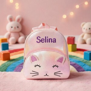May include: A small, iridescent pink backpack featuring a cat face design with glittery ears, embroidered closed eyes, and whiskers. The name Selina is embroidered in purple thread on the upper section of the bag, which includes a top handle and zipper.