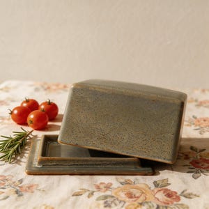 May include: A rectangular, ceramic butter dish in a muted green-grey hue, with a matching lid. The dish is open, revealing the interior. Cherry tomatoes and rosemary sprigs are arranged nearby on a floral patterned tablecloth. The dish is designed for kitchen use.