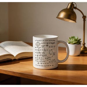 Puede incluir: Taza de cerámica blanca con ecuaciones matemáticas negras y el texto "The answer to Life, universe, and everything." La taza está sobre una superficie de madera, cerca de un libro abierto, una lámpara de escritorio y una pequeña suculenta en maceta.