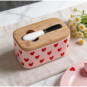 May include: A pink ceramic butter dish with a bamboo lid and a matching butter knife. The dish is decorated with red hearts. A small bouquet of white and yellow flowers is in the background.