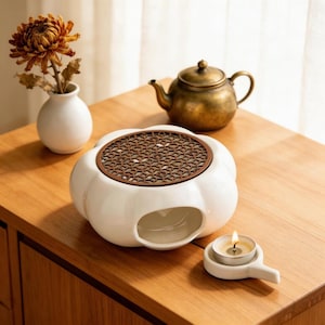 May include: A white ceramic tea warmer with a brown patterned top and a small opening for a tea light candle. A small brass teapot and a white vase with a dried flower are also on the wooden surface.