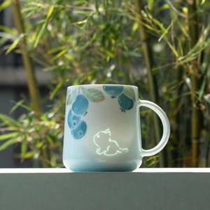 Op de afbeelding: Een keramische mok met een lichtblauwe basis en handvat. De mok heeft een ontwerp van blauwe bessen en bladeren in tinten blauw en groen. Een gloeiend katten silhouet staat ook op de mok.