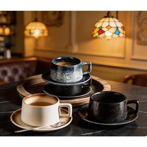Op de afbeelding: Een verzameling keramische koffiekopjes en schotels in verschillende kleuren, waaronder wit, zwart en blauw, op een tafel. De kopjes hebben een getextureerd uiterlijk en zijn gevuld met koffie. Een lepel ligt op de schotel van het witte kopje.