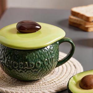 May include: A ceramic avocado-shaped mug with a green exterior and a light green lid. The lid is topped with a brown seed-shaped handle. The mug has a handle and the words "AVOCADO CUP" are embossed on the side.