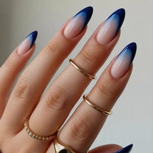 Pode incluir: Close-up de uma mão com unhas longas e ovais. As unhas apresentam um design gradiente, que transita de uma base clara para um azul profundo nas pontas. Vários anéis dourados adornam os dedos, adicionando um toque de elegância.