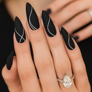Puede incluir: Uñas de estilete negras mate con diseños de líneas blancas curvas. Una mano muestra la manicura, con un anillo de diamantes en forma de pera en el dedo anular. Las uñas son largas y almendradas.