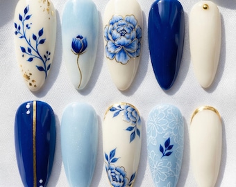 May include: A set of ten artificial nails with various designs. The nails feature a mix of solid colors, floral patterns, and gold accents. Colors include white, light blue, and navy blue. The floral designs are in blue.