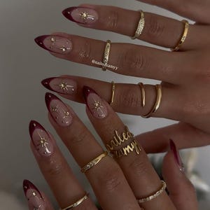 Op de afbeelding: Close-up van gemanicuurde nagels met een bordeauxrode French tip en gouden steraccenten. De nagels zijn versierd met kleine gouden sterren en blanke nagellak. Verschillende gouden ringen van verschillende ontwerpen worden op de vingers gedragen.