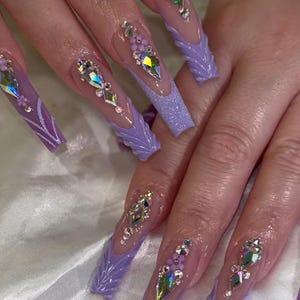 May include: Long, lavender-colored acrylic nails with intricate designs. Each nail features a clear base, embellished with sparkling rhinestones and a glittery lavender tip. Some nails have a textured, leaf-like design.